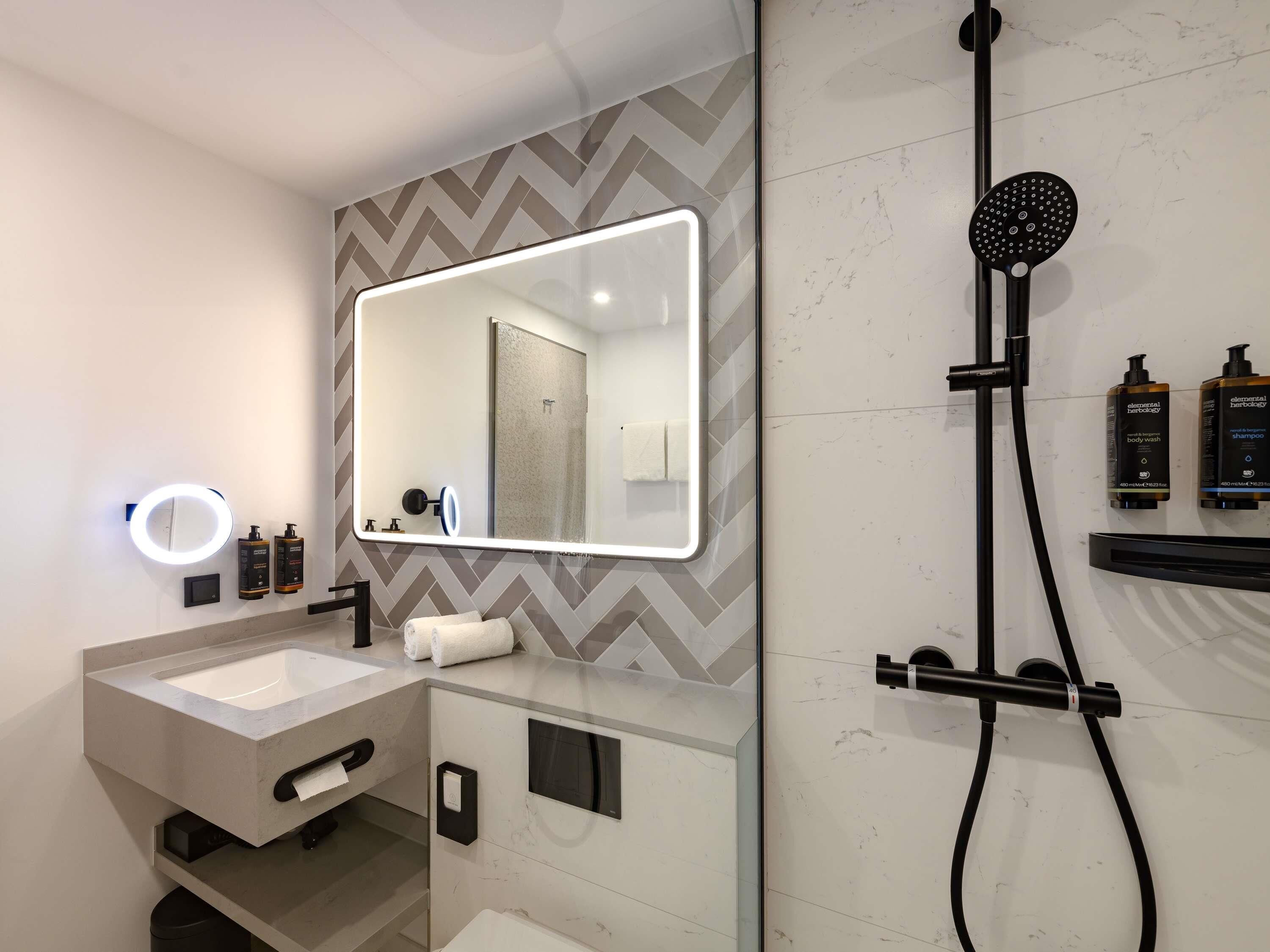 A modern bathroom with a large mirror, a handheld showerhead, a vanity with a sink, and two bottles of shampoo.