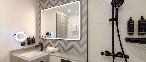 A modern bathroom with a large mirror, a handheld showerhead, a vanity with a sink, and two bottles of shampoo.