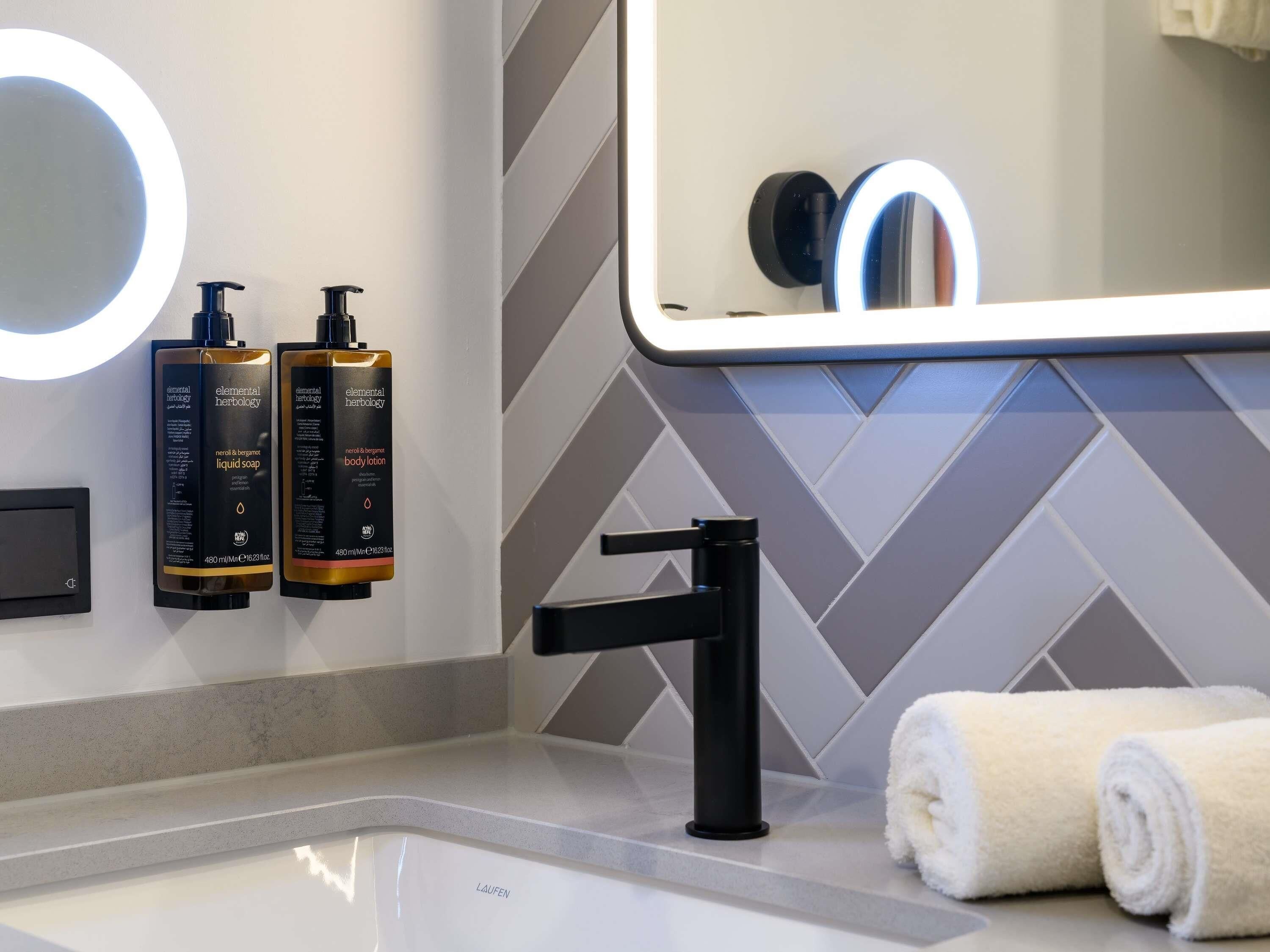 A bathroom with a modern design, featuring a large mirror, two bottles of Elemental Herbology body lotion, a black faucet, and a towel on the counter.
