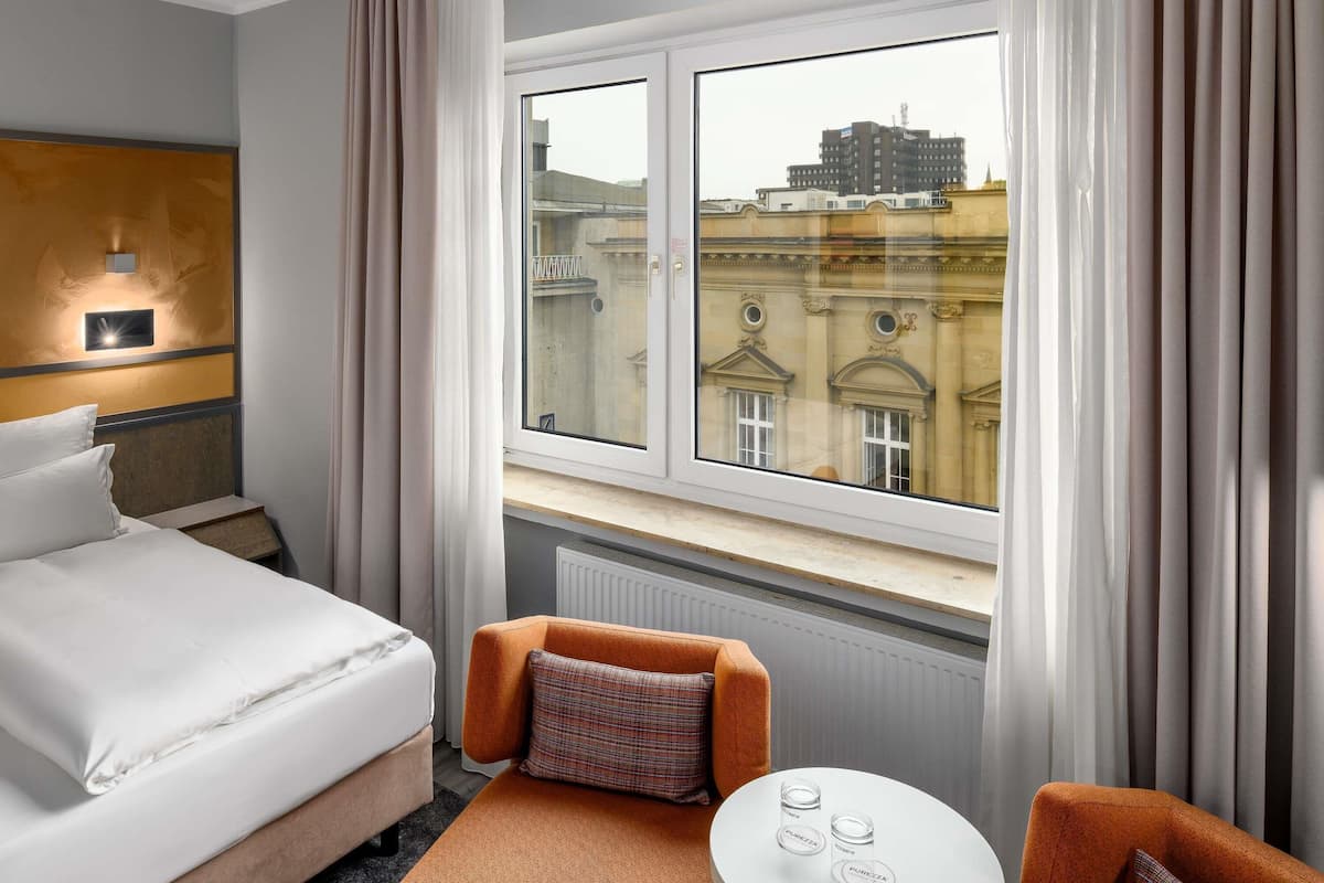 Une chambre d’hôtel avec un lit, un canapé, une table avec des verres et une fenêtre donnant sur des immeubles.