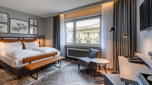 Habitación de hotel con cama, escritorio, silla, ventana con vistas a edificios y cuadros en la pared.