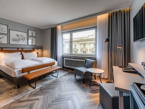 Une chambre dâhĂŽtel avec un lit, un bureau, une chaise, une fenĂȘtre donnant sur des immeubles et des tableaux encadrĂ©s accrochĂ©s au mur.