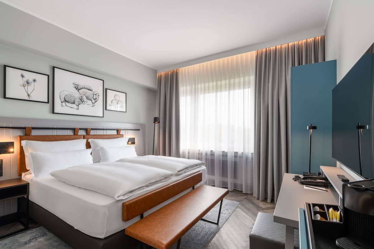 Une chambre d’hôtel moderne dotée d’un grand lit, de tables de chevet, d’un bureau avec un téléviseur à écran plat et d’une grande fenêtre avec des rideaux.