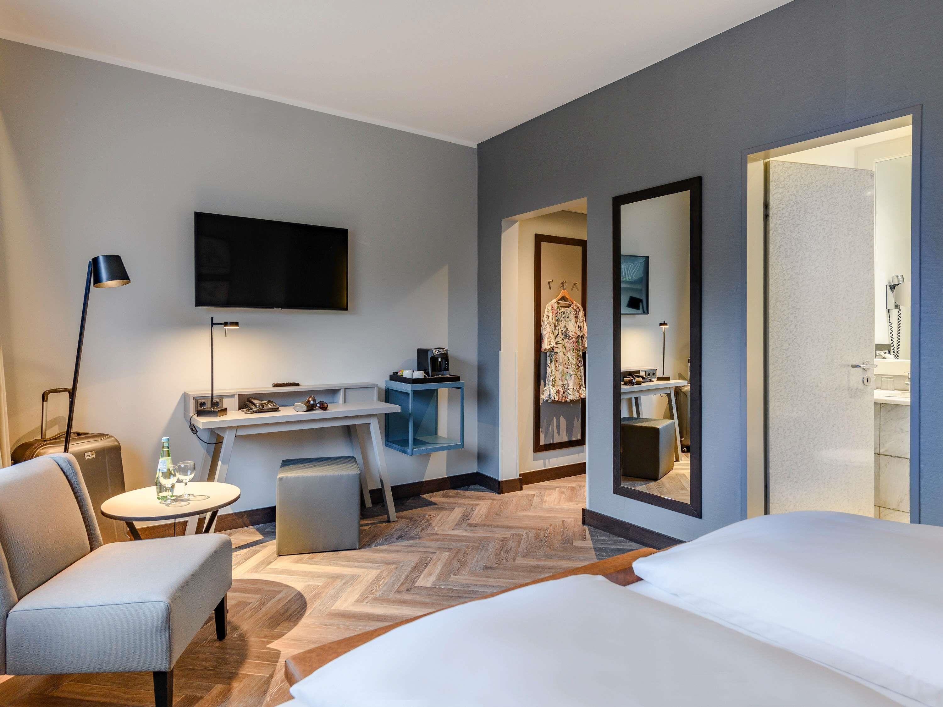 A hotel room with a bed, a desk, a chair, a TV, a mirror, and a bathroom visible through an open door.
