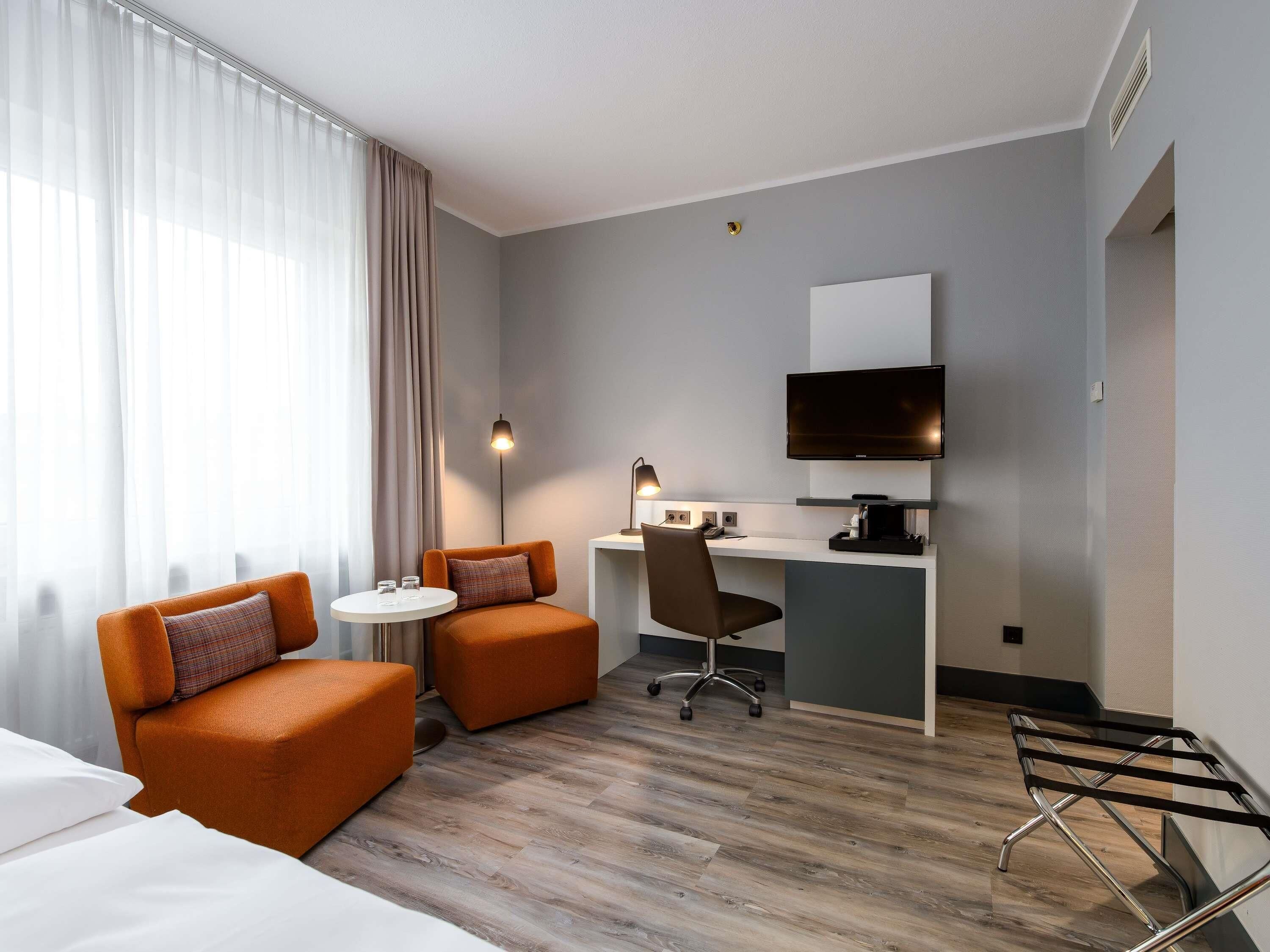 Una camera d'albergo con un letto, due poltrone arancioni, una scrivania con un computer e una TV, e un tavolino con una lampada.