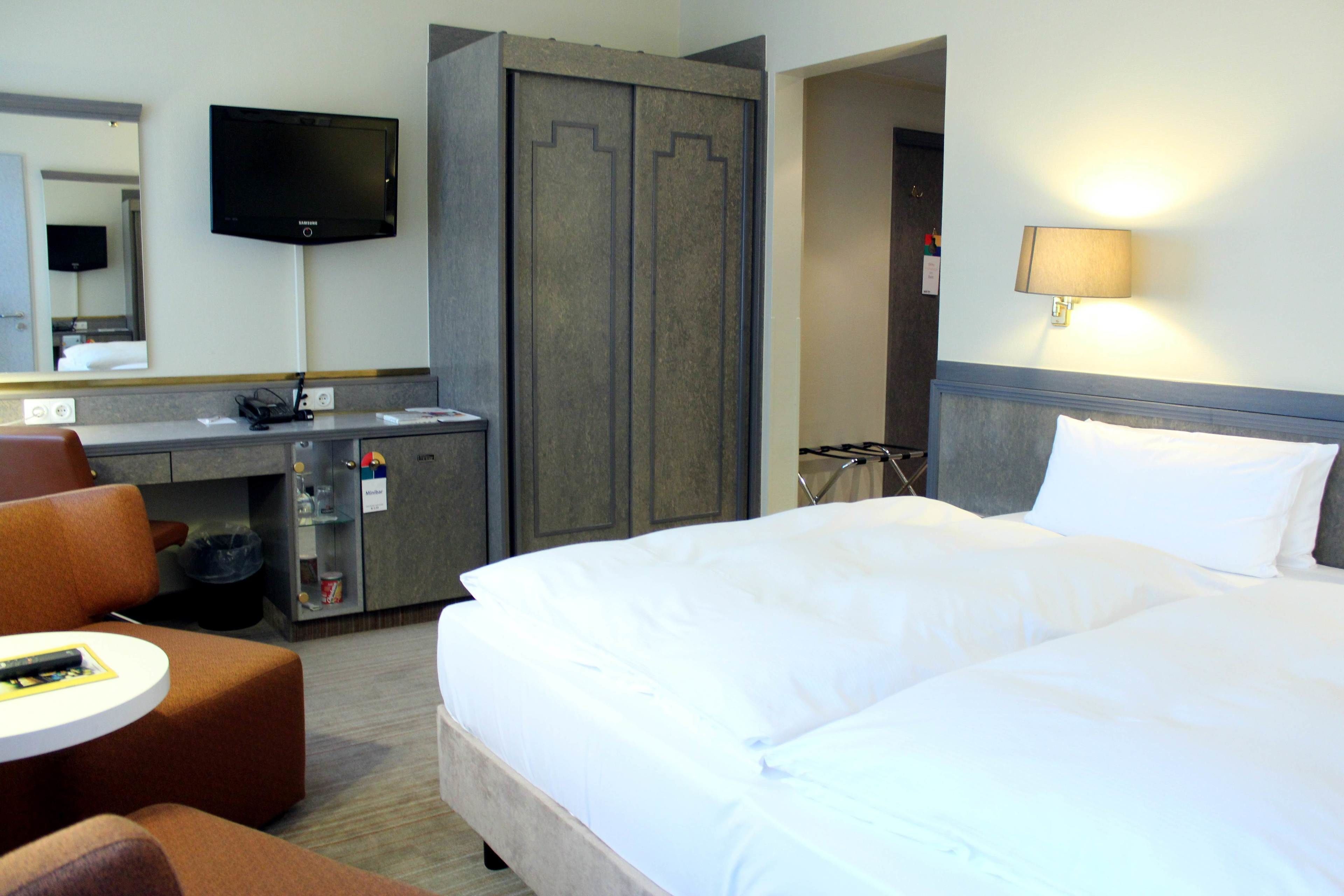 A hotel room with a bed, a desk with a chair, a television, and a wardrobe.