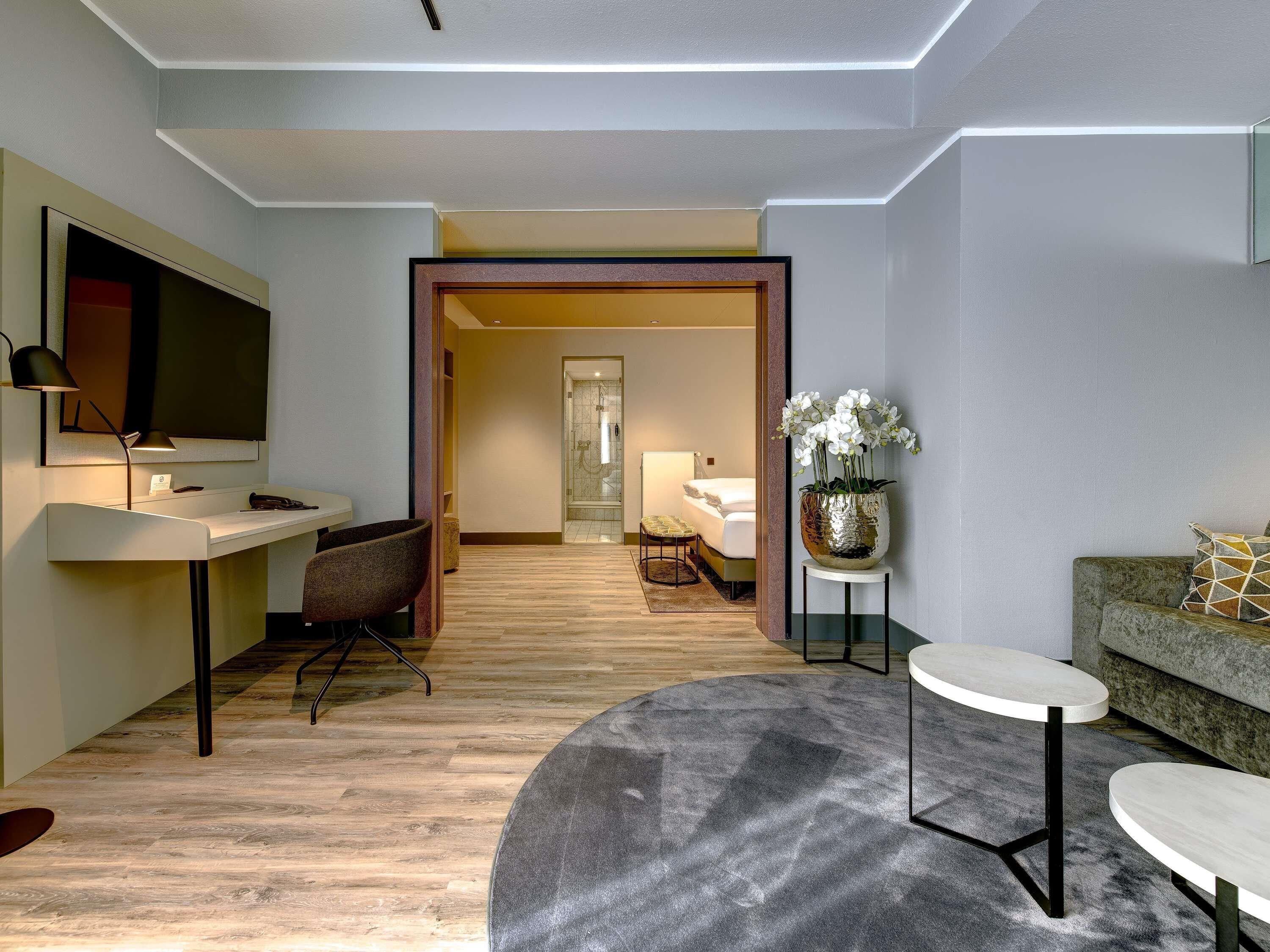 Habitación de hotel moderna con escritorio, silla, sofá y una pequeña mesa. Se ve una zona de ducha al fondo.