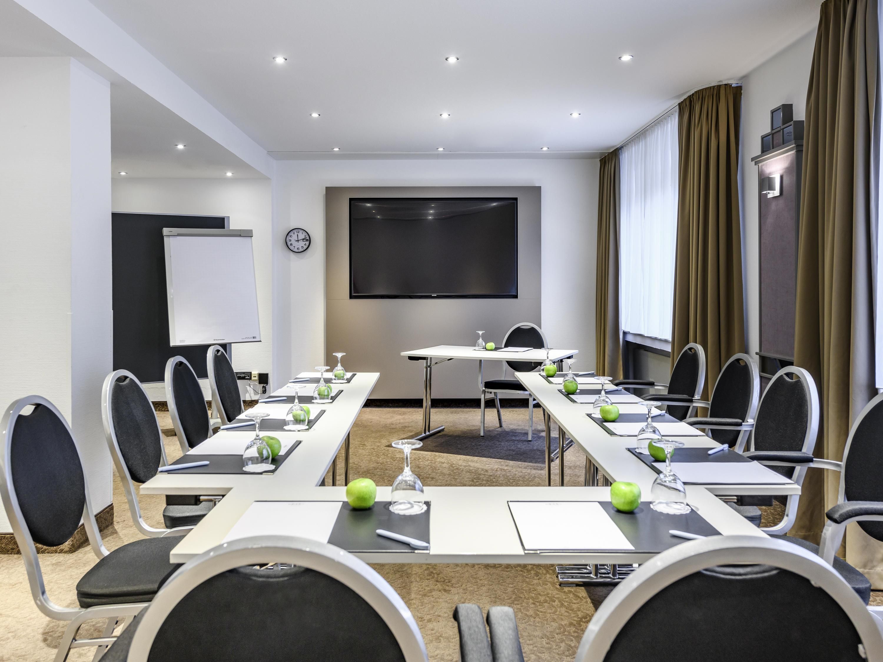 Une salle de conférence avec une longue table, des chaises, un écran de projection et une horloge au mur.