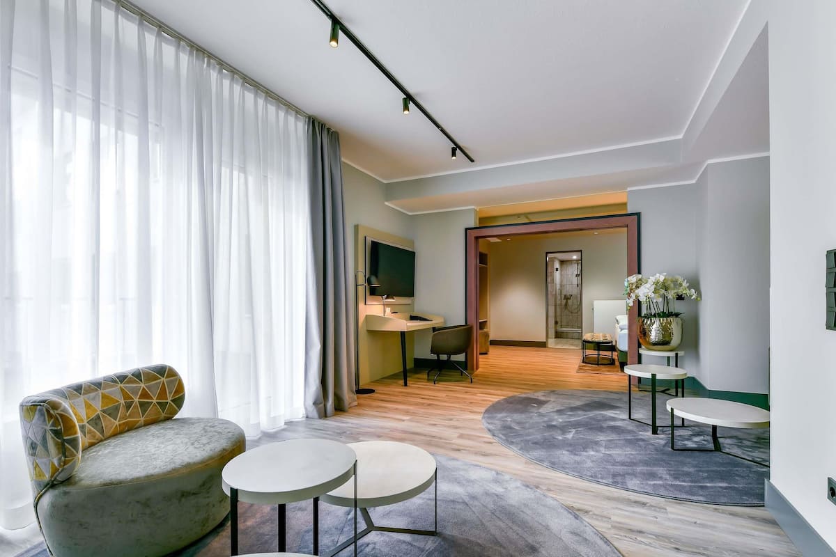 A modern hotel room with a sofa, two small tables, a TV, and a bathroom visible through an open door.
