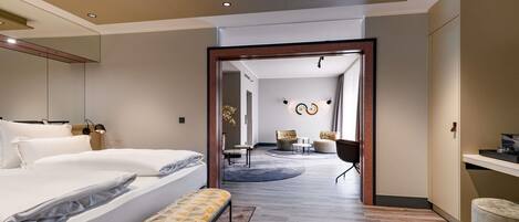 A modern hotel room with a large bed, a bench, a desk, and a seating area with chairs and a table.