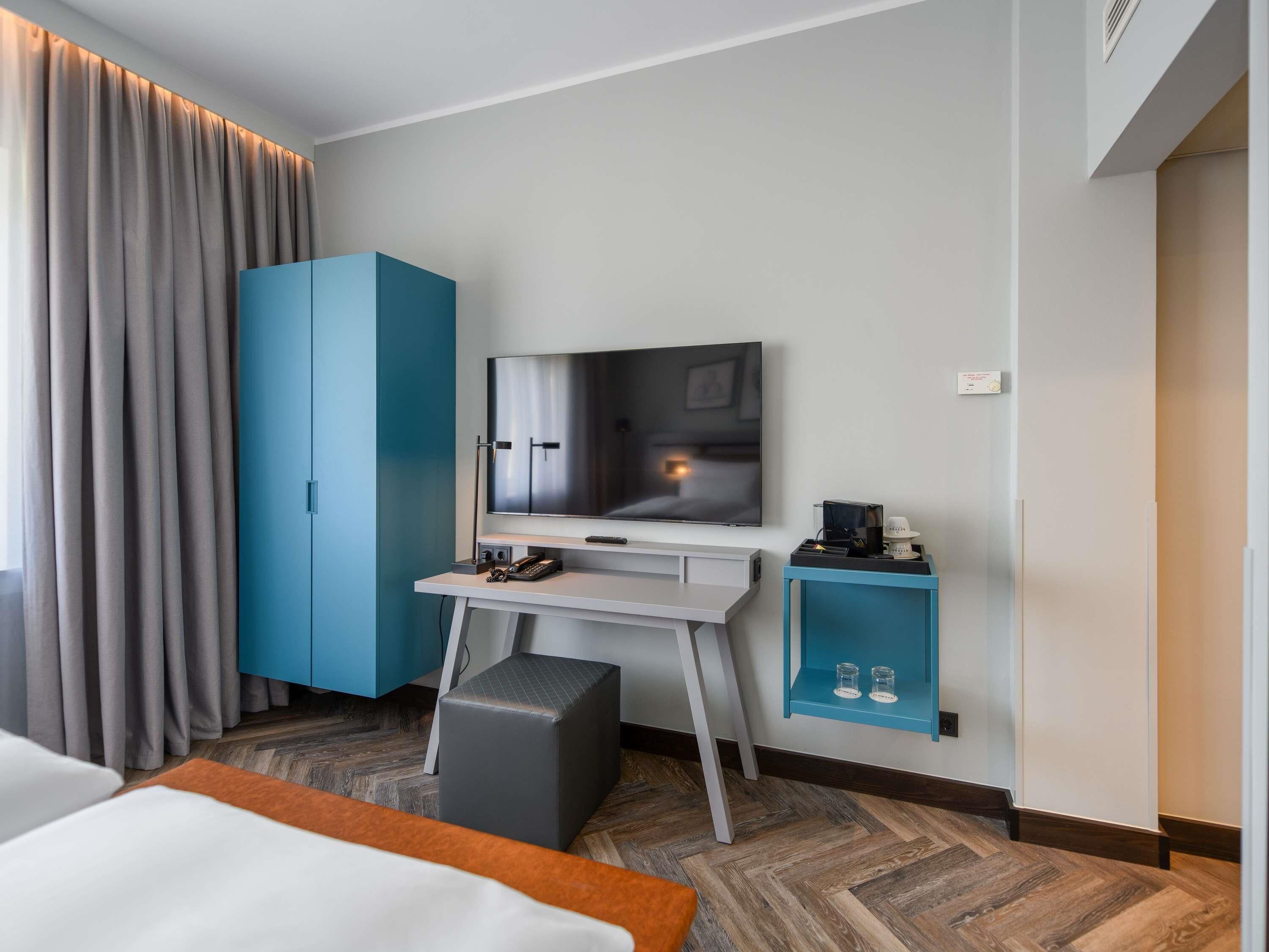 A modern hotel room with a large bed, a desk with a flat-screen TV, a wardrobe, and a small side table with a coffee maker.