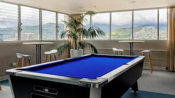 A pool table in a room with large windows, a potted plant, and two small tables with chairs.