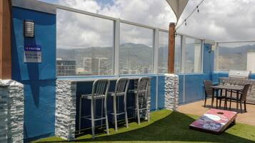 Uma área externa com mesas e cadeiras, vista para a piscina, pilares de pedra e churrasqueira.