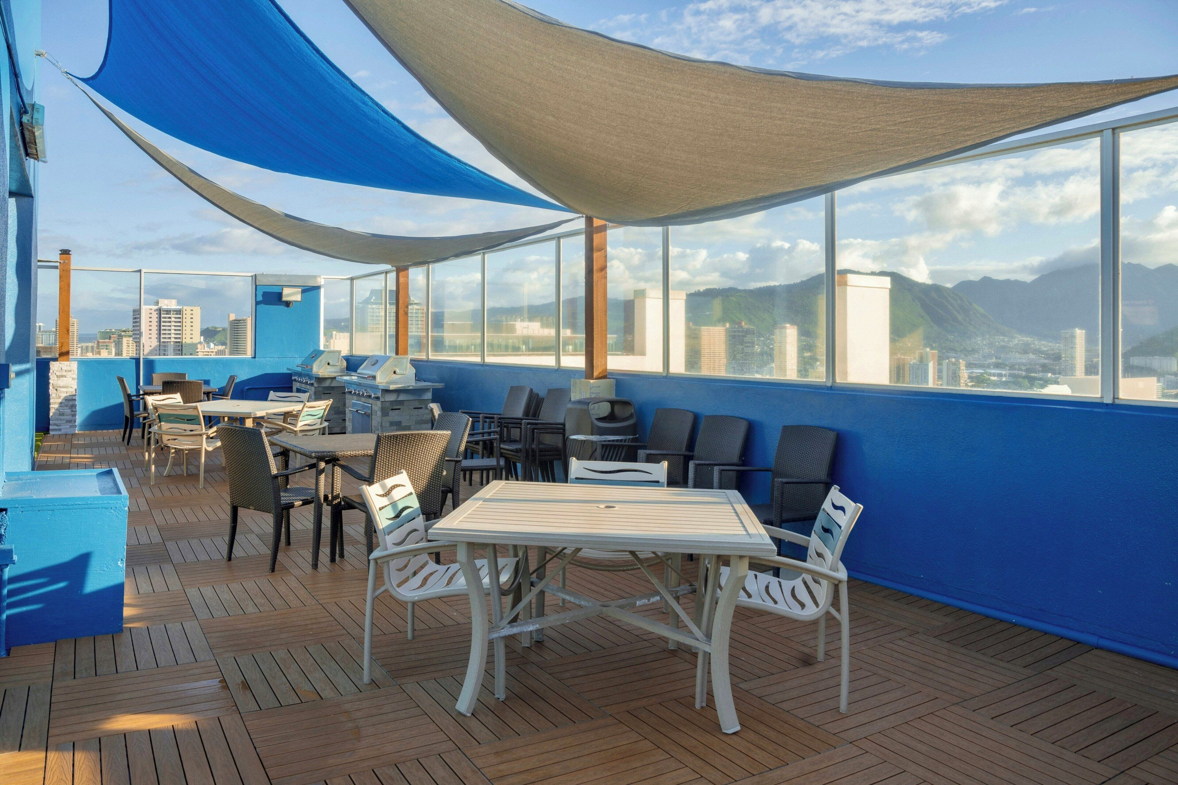 Una terraza exterior con mesas y sillas, una parrilla y vistas a un paisaje urbano y montañas.