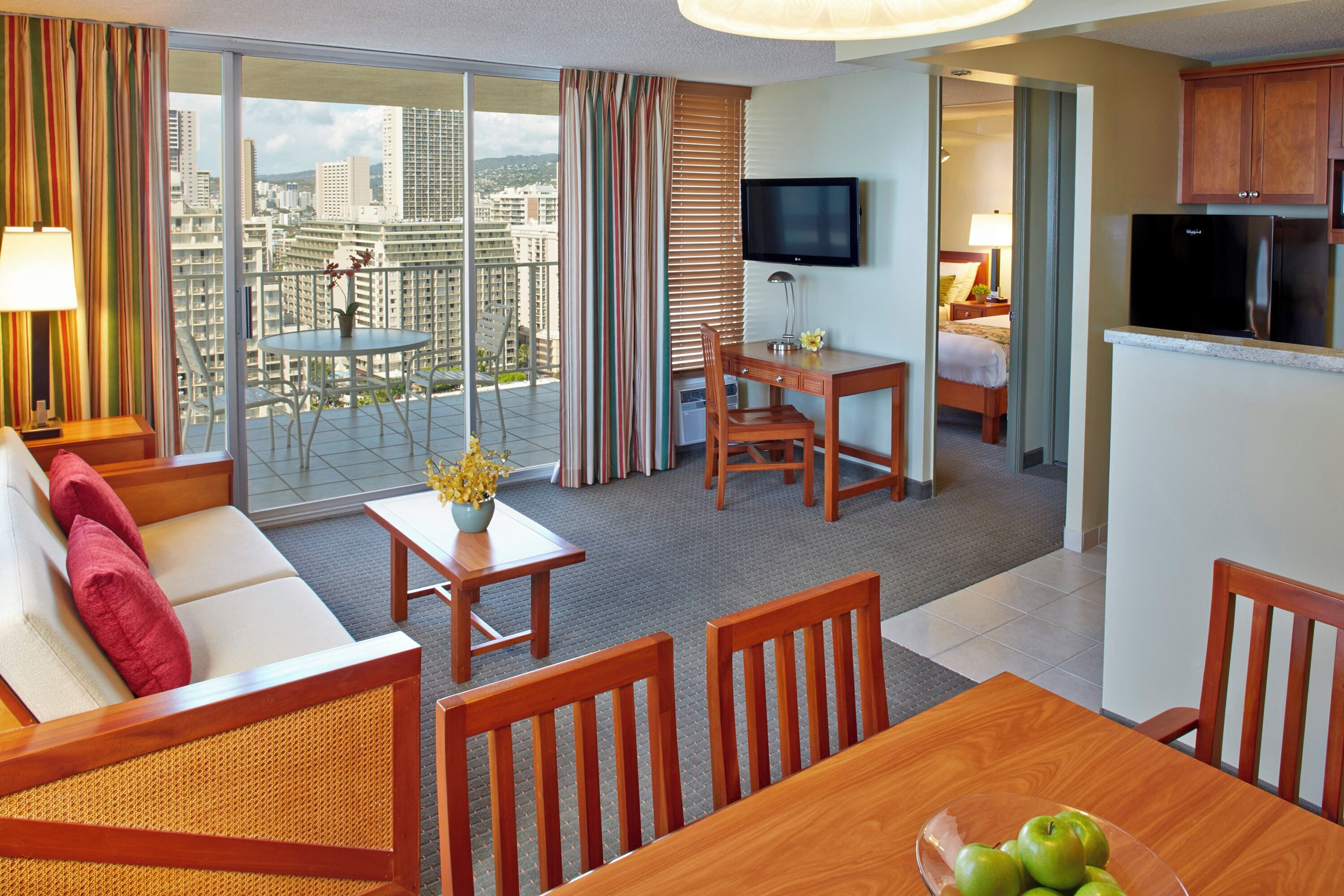 A hotel room with a balcony, a dining area, a sofa, a TV, and a kitchen area.