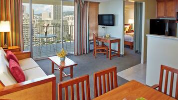 A hotel room with a balcony, a dining area, a sofa, a TV, and a kitchen area.