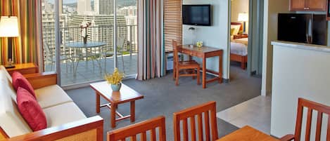 A hotel room with a balcony, a dining area, a sofa, a TV, and a kitchen area.