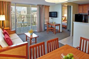 Habitación de hotel con balcón, zona de comedor, sofá, televisor y cocina.