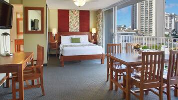 A hotel room with a bed, a desk, a chair, a table, a TV, a ceiling fan, and a view of the city.