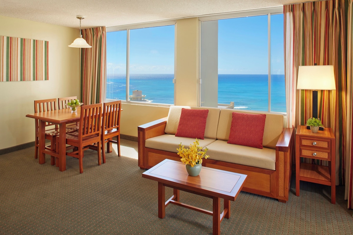 A hotel room with a dining table, sofa, and a view of the ocean.