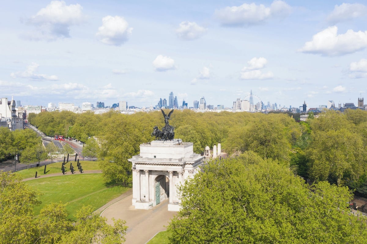 Un parque con un monumento, rodeado de árboles y un paisaje urbano al fondo.