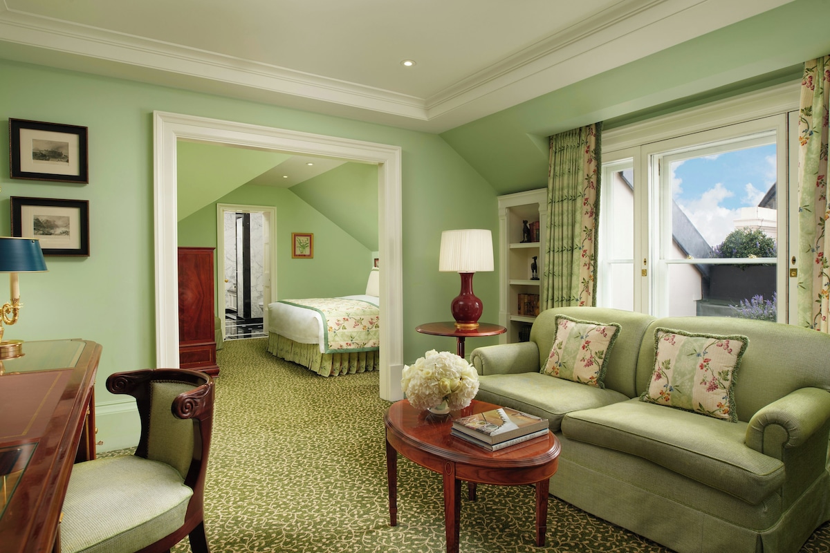 A room with a green carpet, a green sofa with floral cushions, a wooden side table with a vase and book, a wooden chair, a bed with a floral bedspread, and a door leading to another room.