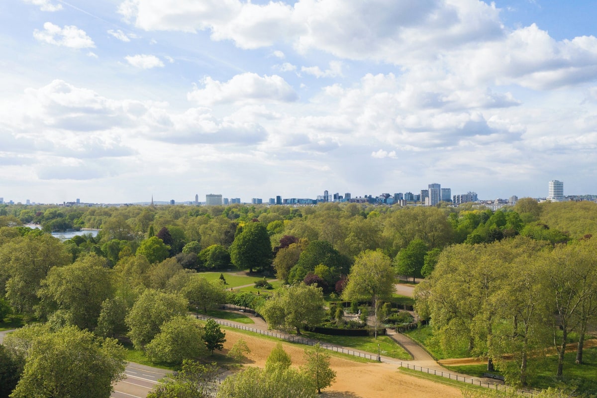 A park with pathways and trees, a body of water, and a distant city skyline.