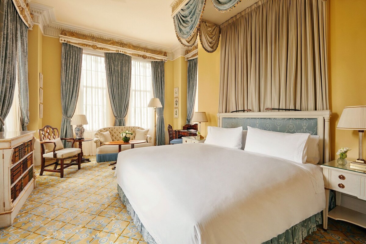A large bed with white linens, a wooden nightstand, a chair, a small table with a lamp, and a bookshelf.