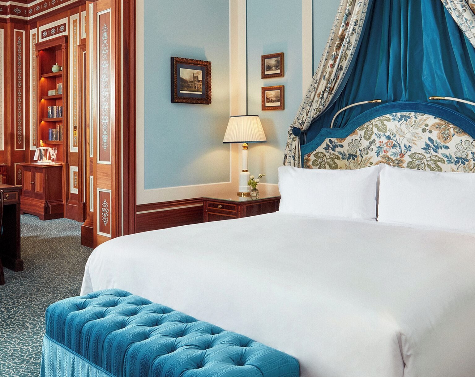 A hotel room with a large bed, a nightstand with a lamp, a wooden wardrobe, and a blue upholstered bench.