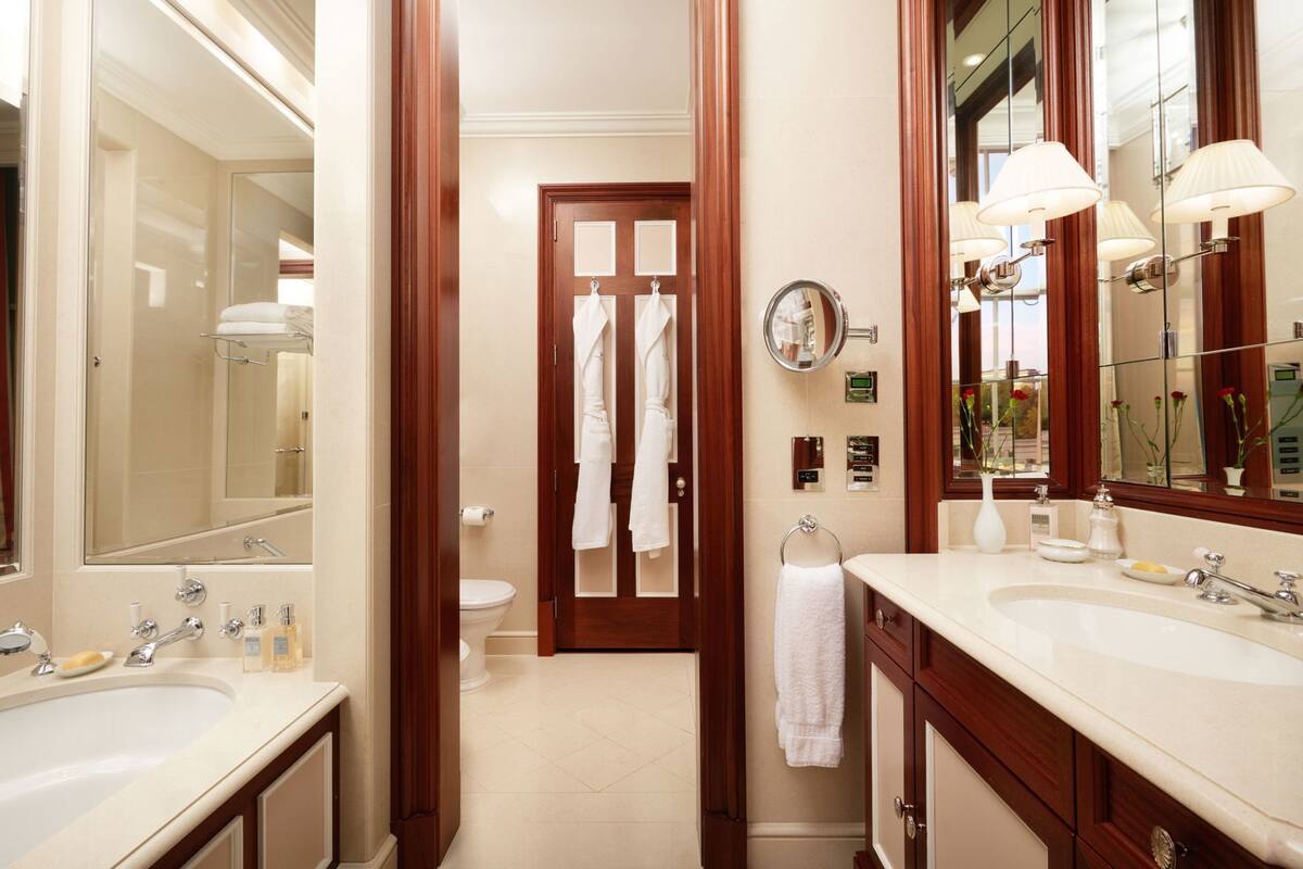 Un baño de hotel con dos lavamanos, un espejo, un perchero para toallas y una puerta que da al inodoro.
