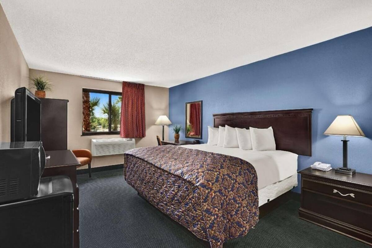 A hotel room with a bed, a desk, a TV, a window with curtains, and a nightstand with a lamp.
