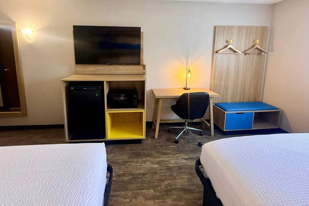 A hotel room with two beds, a desk, a chair, a TV, a wardrobe with hangers, and a small shelf with a lamp.