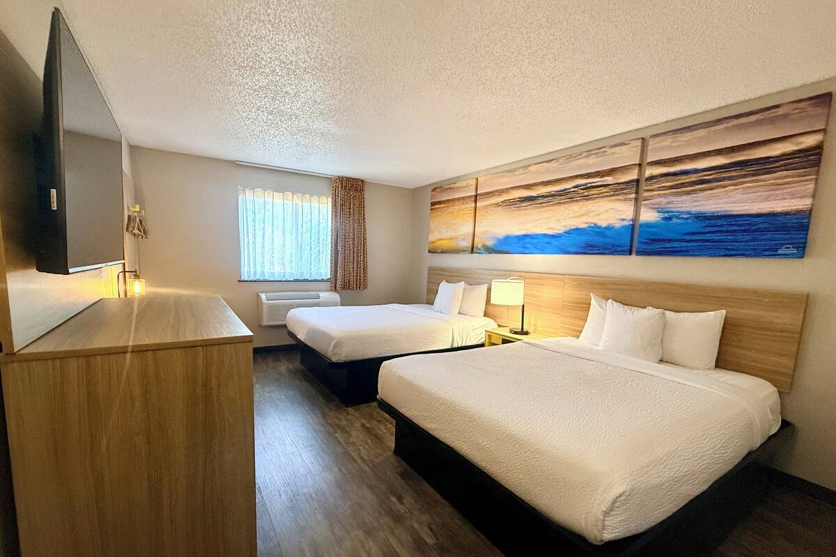 A hotel room with two beds, a desk, a flat-screen TV, a painting, and a window with curtains.