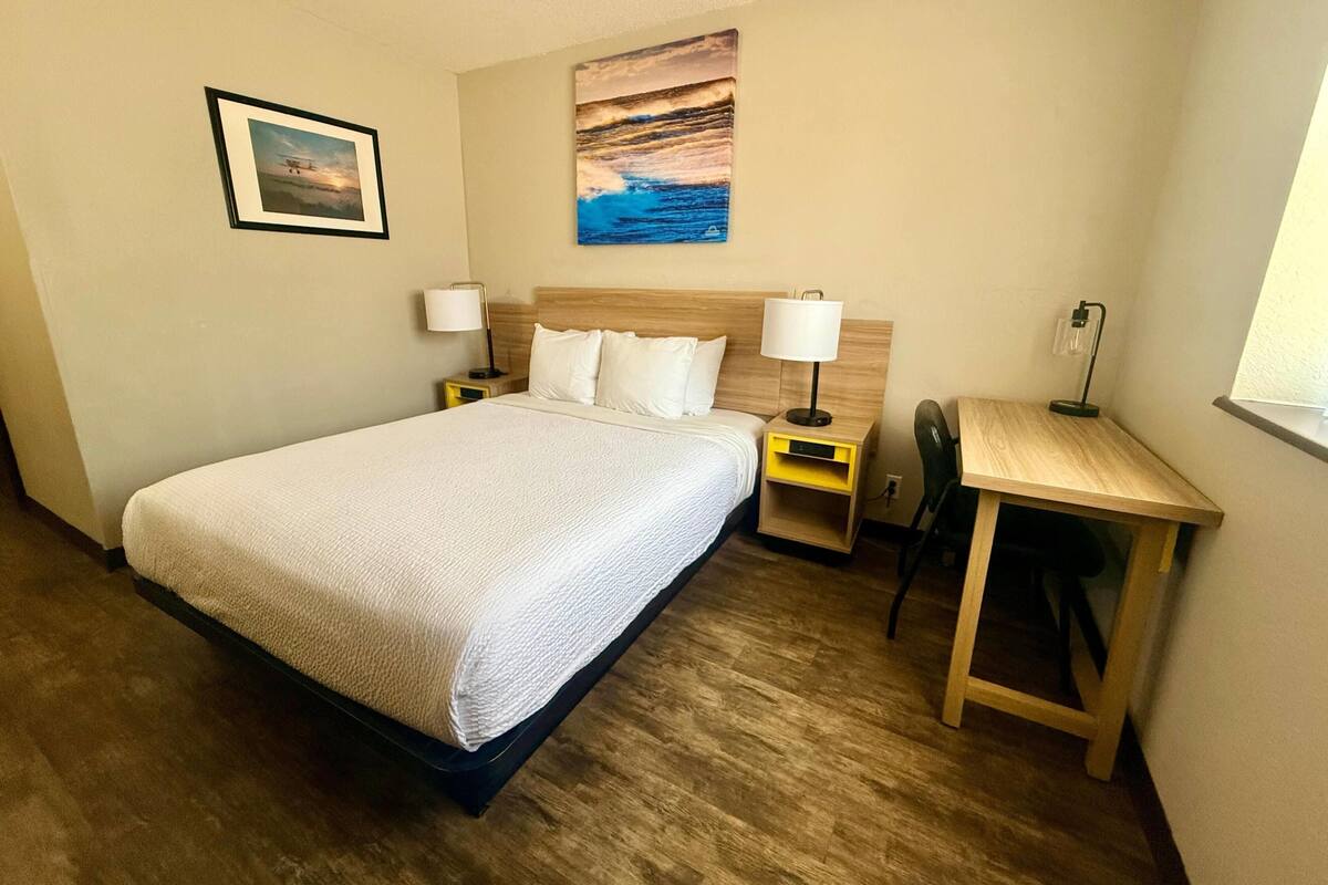 A hotel room with a bed, bedside tables with lamps, a desk, and a painting on the wall.