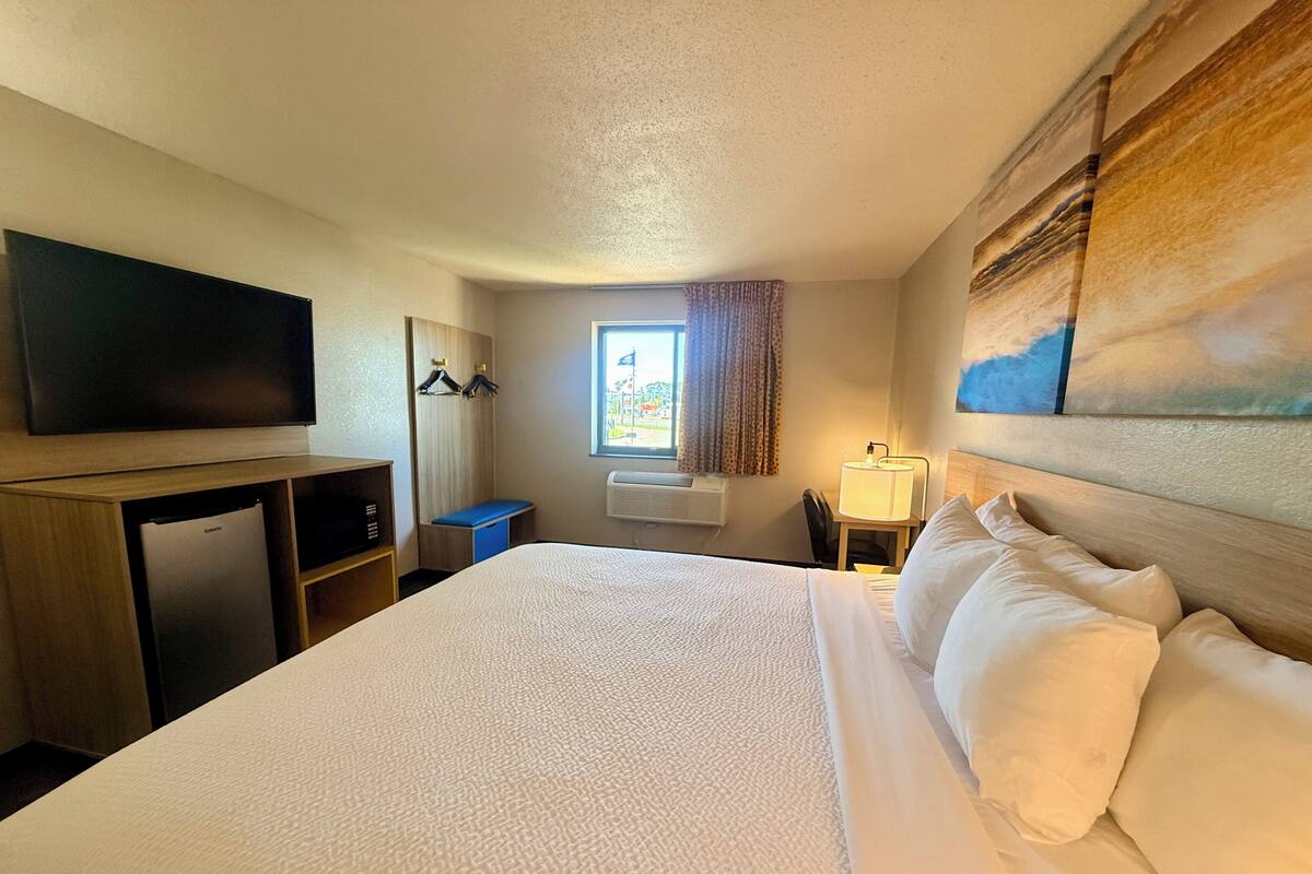 A hotel room with a bed, a flat-screen TV, a desk, a chair, a window with curtains, and a painting on the wall.