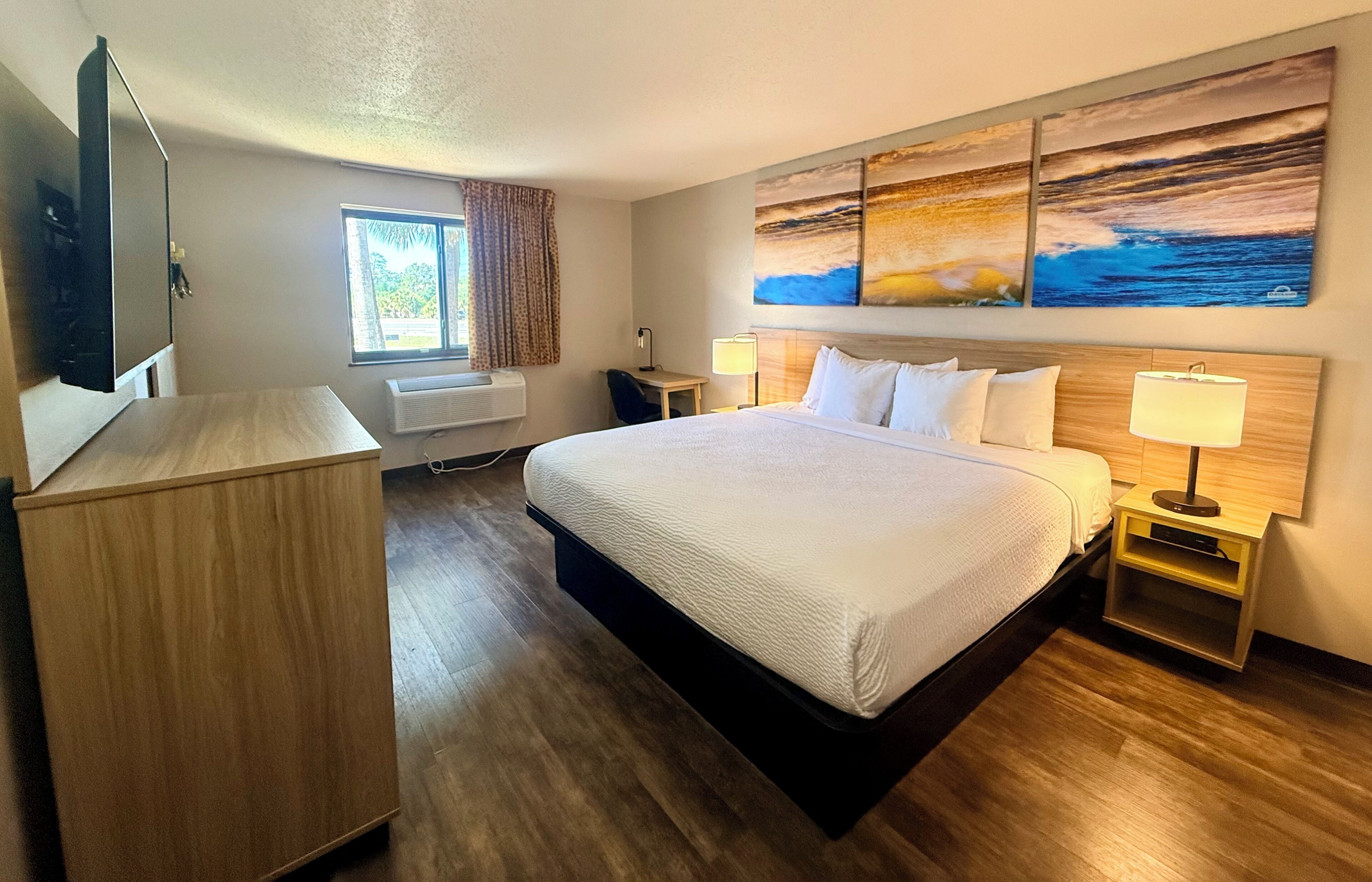 A hotel room with a large bed, a desk, a flat-screen TV, a window with curtains, and artwork on the wall.