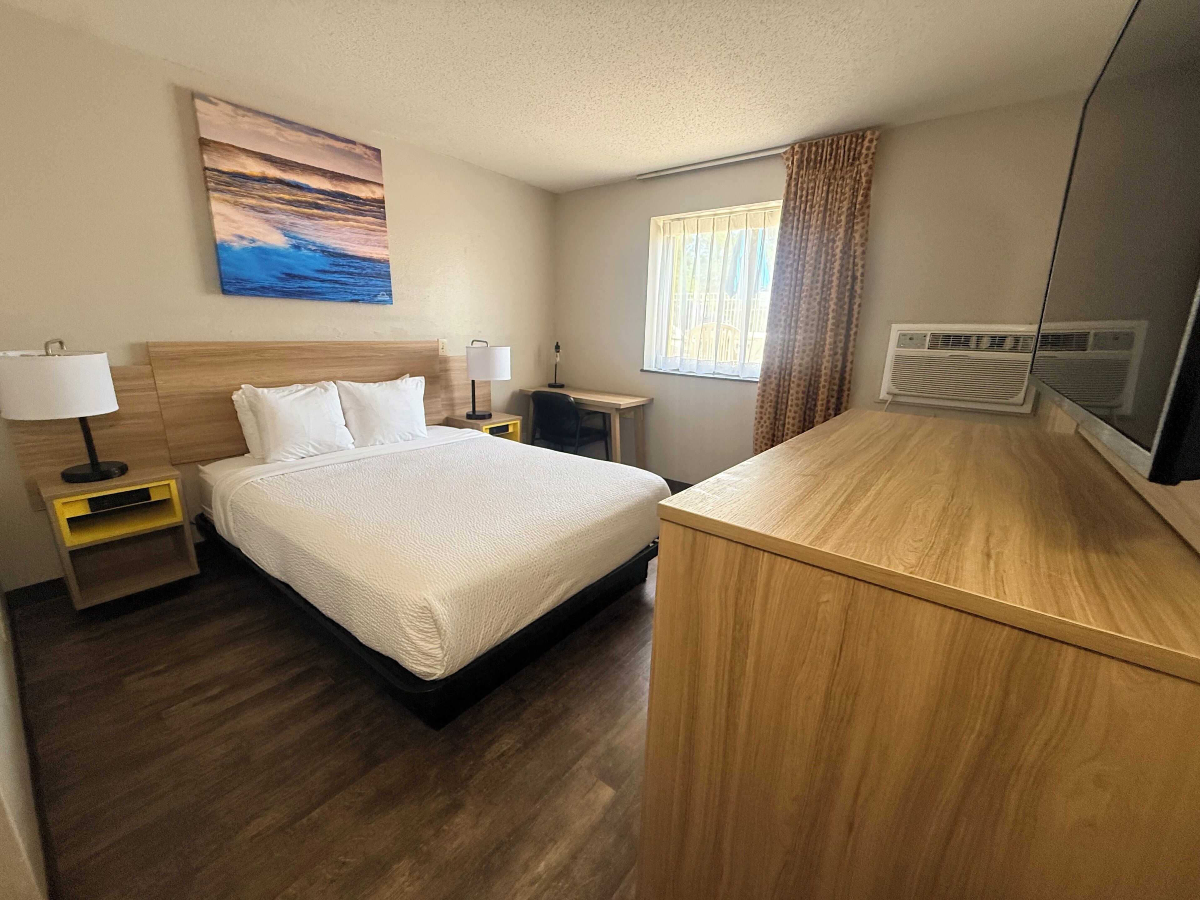Hotelkamer met een bed, nachtkastjes, een bureau, een stoel, een raam met gordijnen, een wandairconditioner en een schilderij.