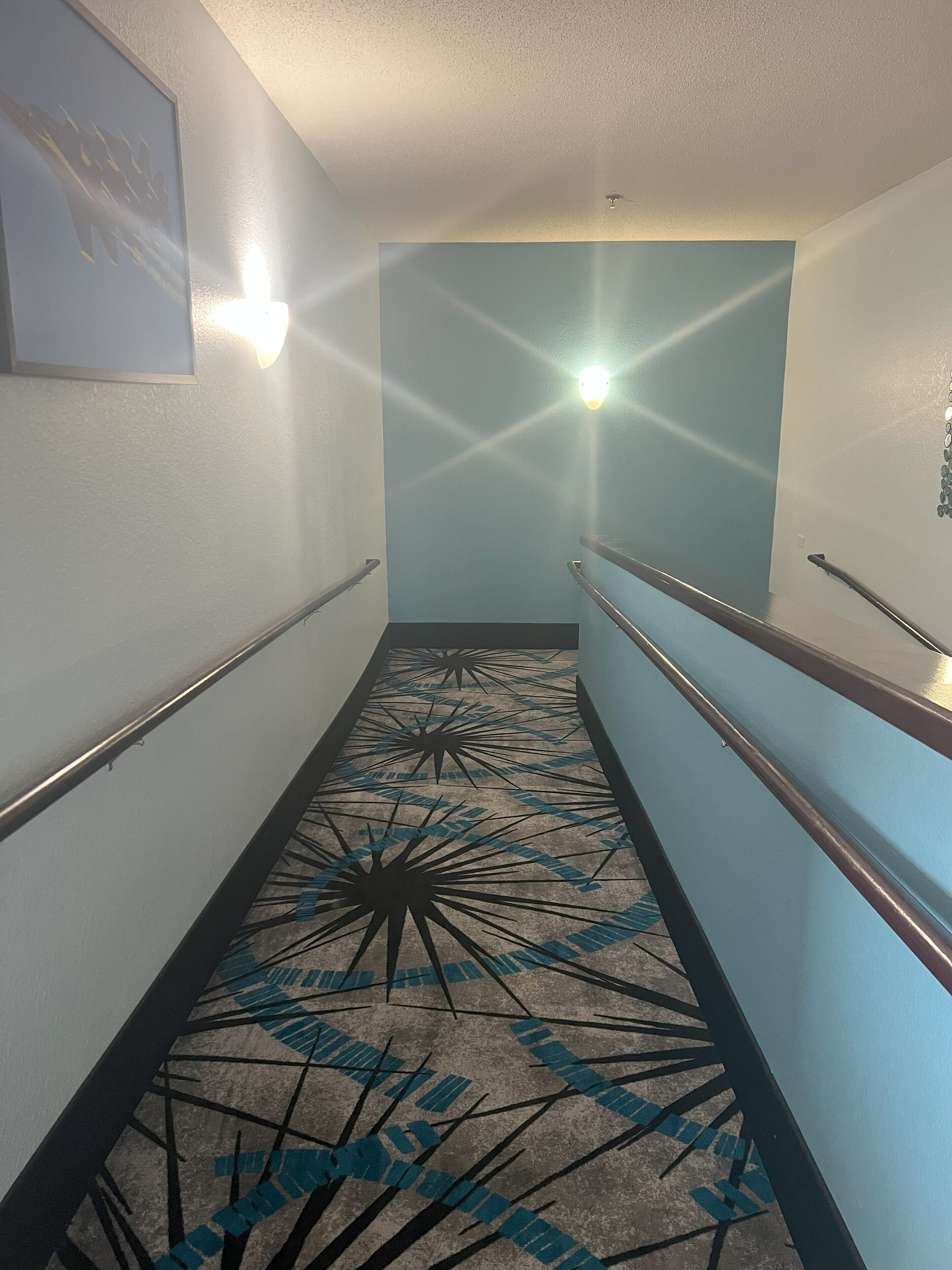 A hallway with a patterned floor, handrails, and wall-mounted lights.