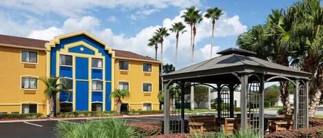A yellow building with a blue entrance, a gazebo with benches, and palm trees in the background.