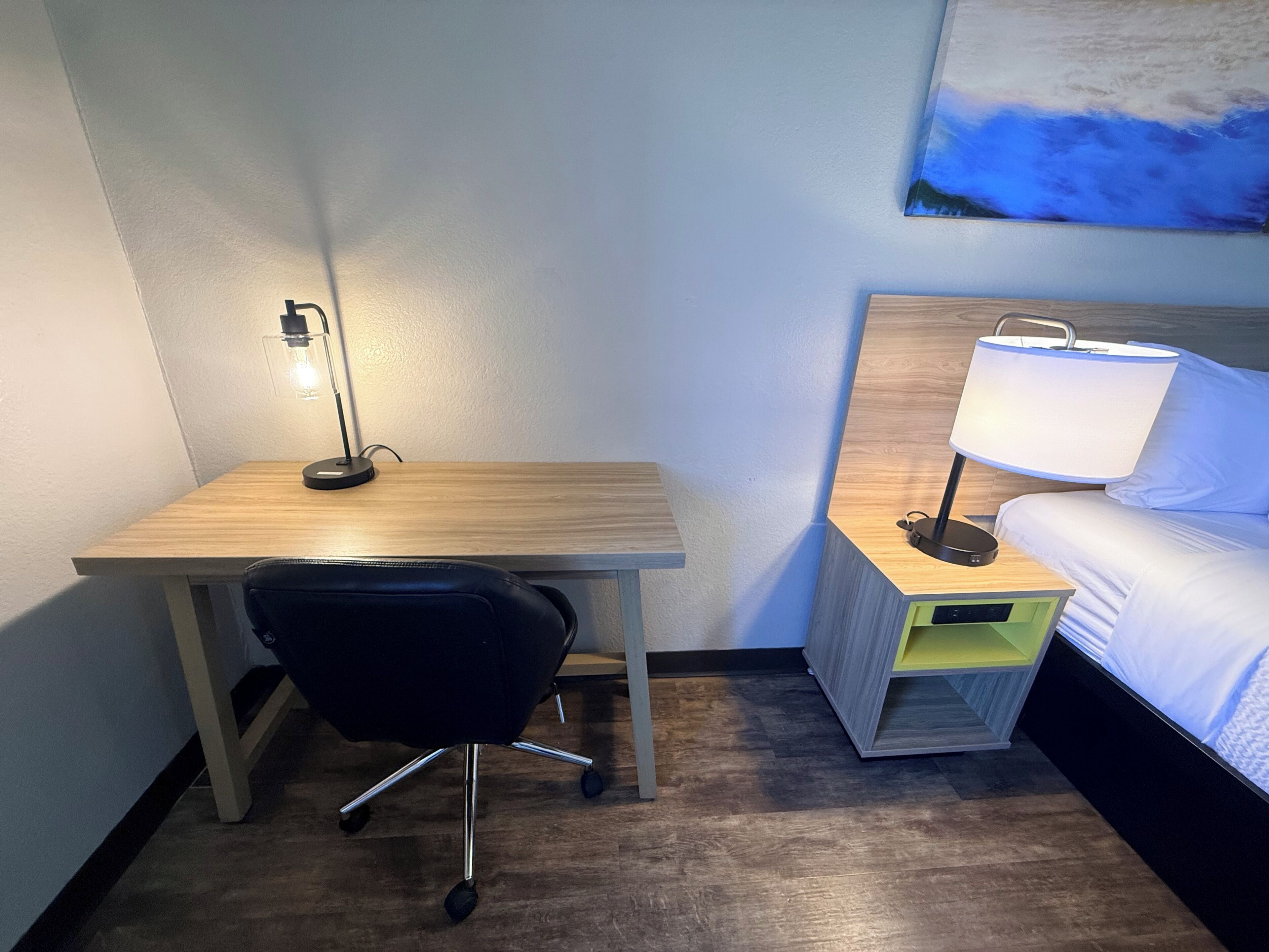 A hotel room with a wooden desk, a black office chair, a bedside table with a lamp, a bed with white bedding, and a painting on the wall.