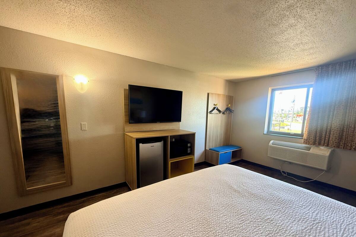 A hotel room with a bed, a television, a mini-fridge, a heater, a window with curtains, and a coat rack.