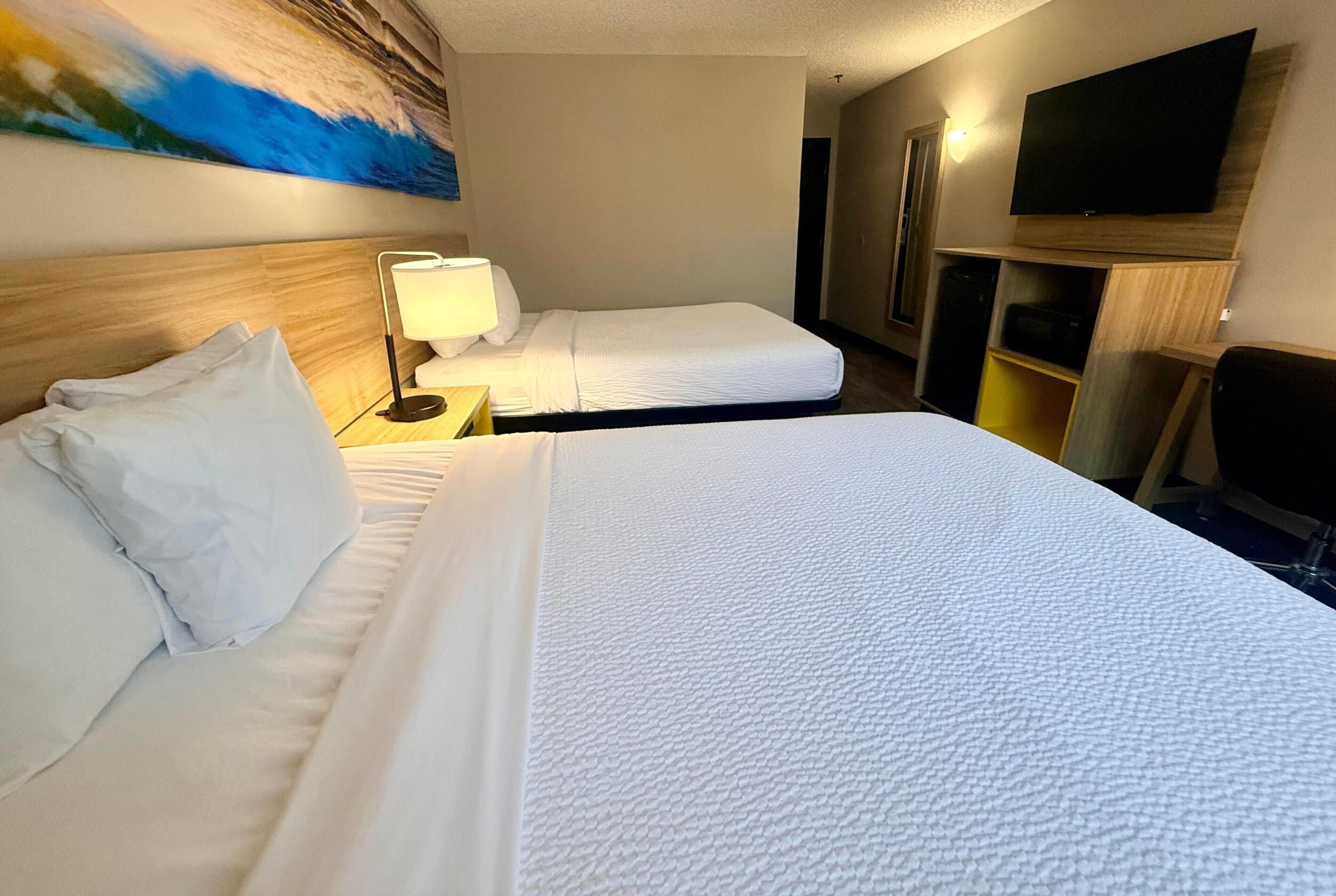 Ein Hotelzimmer mit zwei Betten, einem Fernseher, einem Bild an der Wand und einem Schreibtisch mit Lampe.