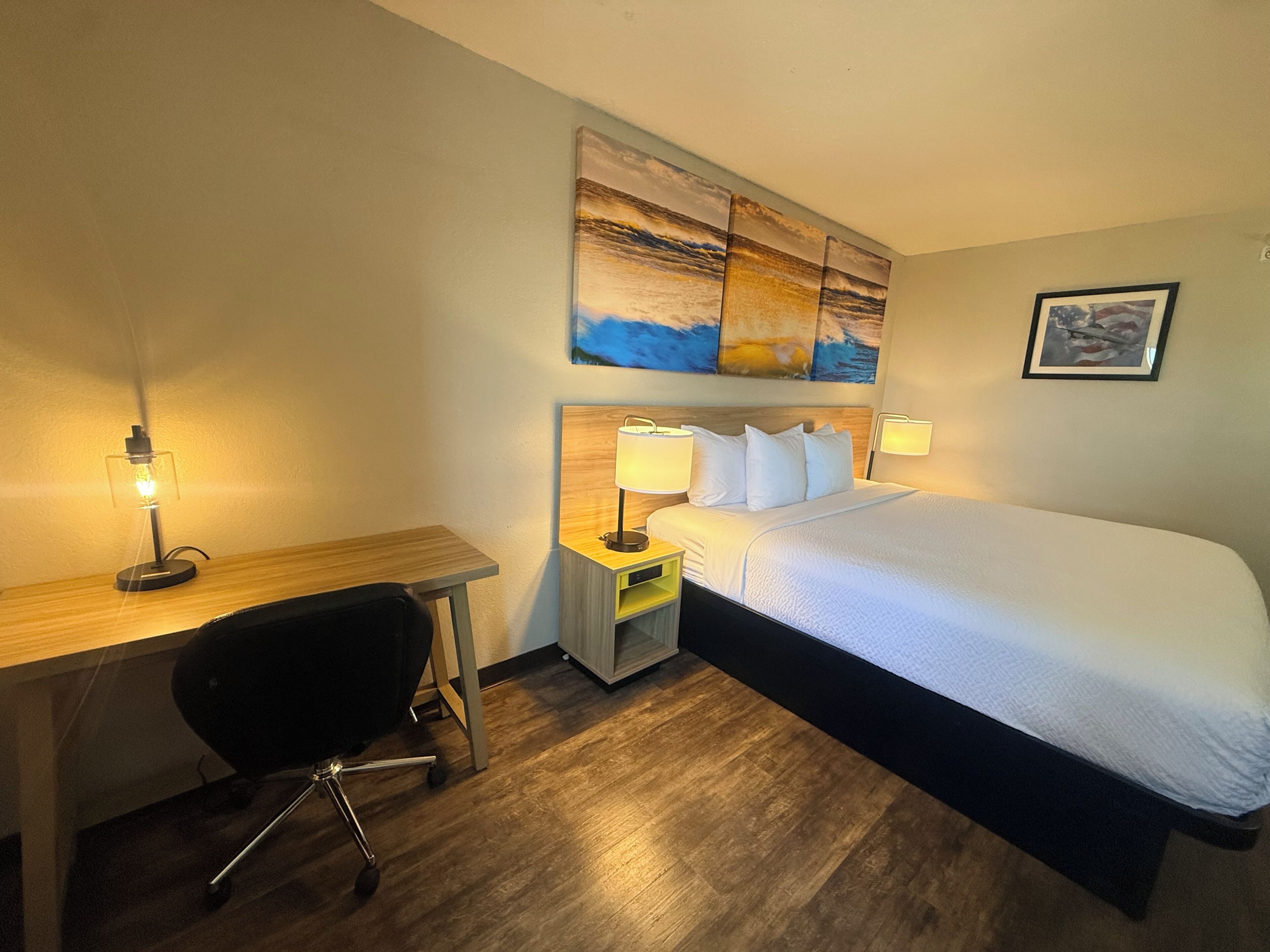 A hotel room with a bed, desk, chair, bedside lamps, and artwork on the wall.