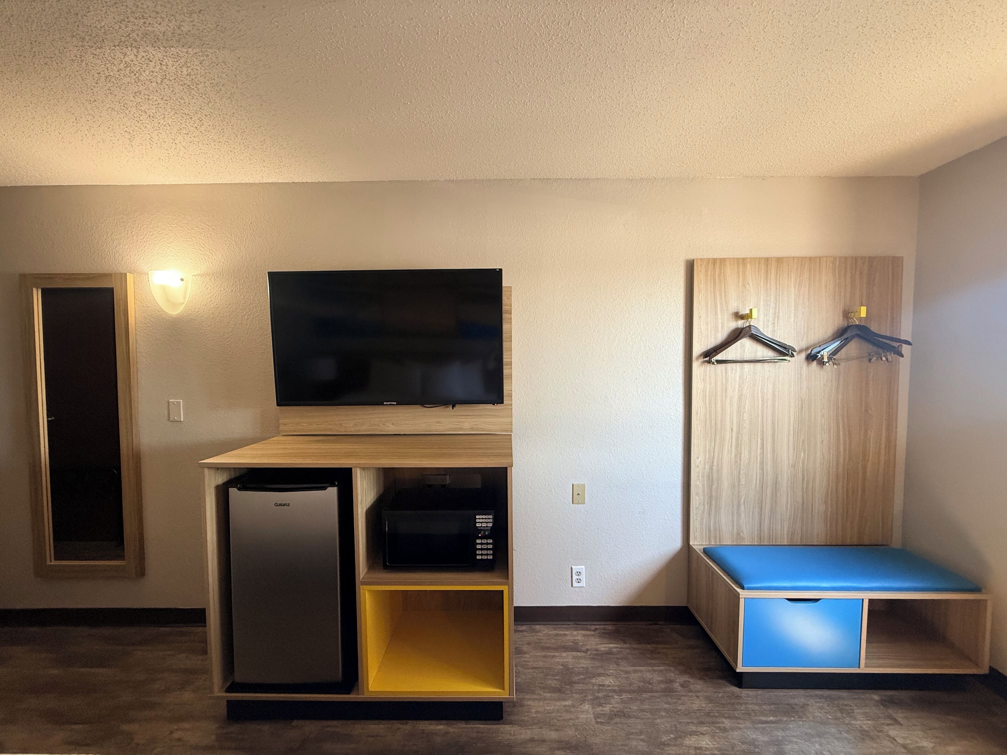A compact room with a flat-screen TV, a built-in mini-fridge, a microwave, a bench, and a wooden panel with two hangers.