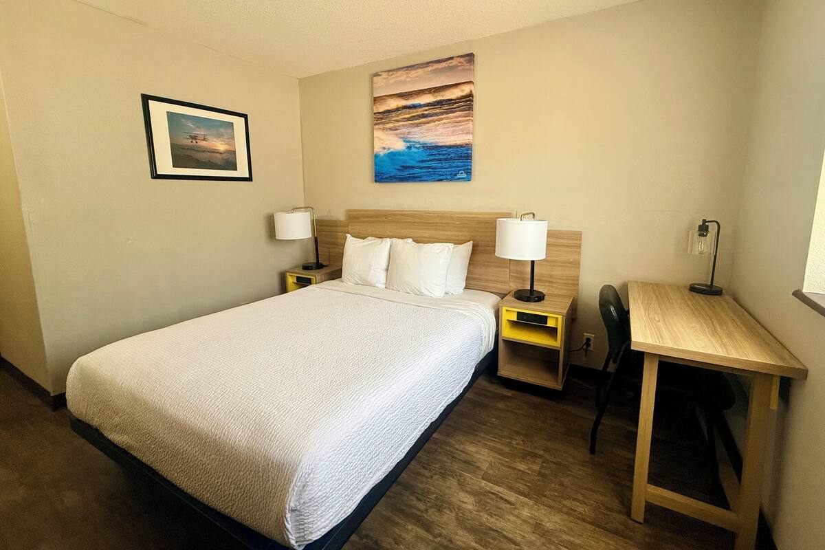 A hotel room with a bed, two bedside tables with lamps, a desk, a chair, and a painting on the wall.