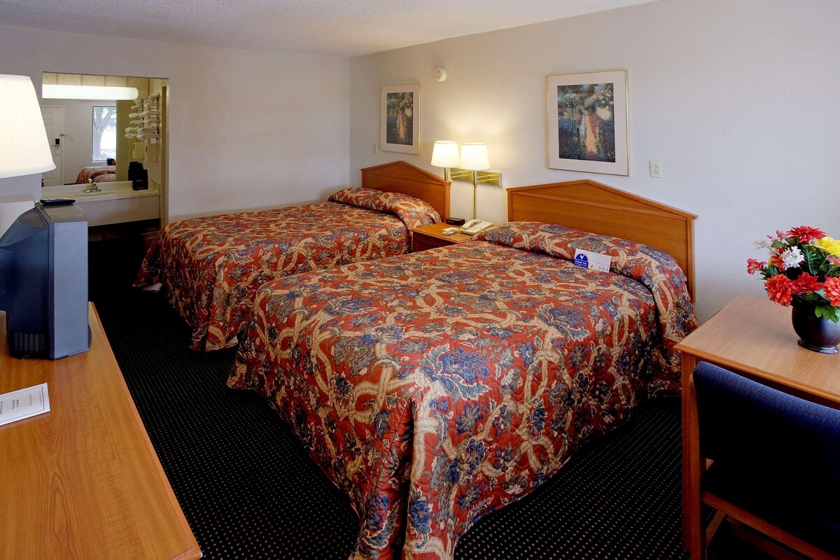 Quarto de hotel com duas camas, uma escrivaninha, uma cadeira, um abajur, um telefone e um vaso com flores.