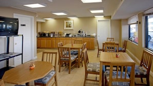 A dining area with wooden tables and chairs, a television on a stand, and a kitchen in the background. - Americas Best Value Inn & Suites Murfreesboro (Murfreesboro)