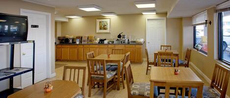 A dining area with wooden tables and chairs, a television on a stand, and a kitchen in the background.