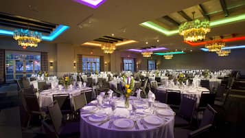Un salón de banquetes con mesas redondas dispuestas para comer, candelabros y grandes ventanales.