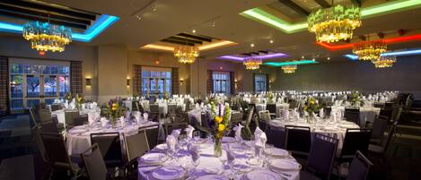 Une salle de banquet avec des tables rondes dressées pour le repas, des lustres et de grandes fenêtres.